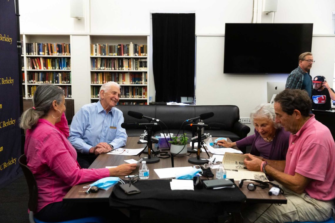 A group of elderly people sit around a table in a library, engaged in conversation and sharing papers. Microphones are positioned in front of them, suggesting a recorded discussion or interview for Berkeley Journalism. Behind them are bookshelves filled with books and a television screen.