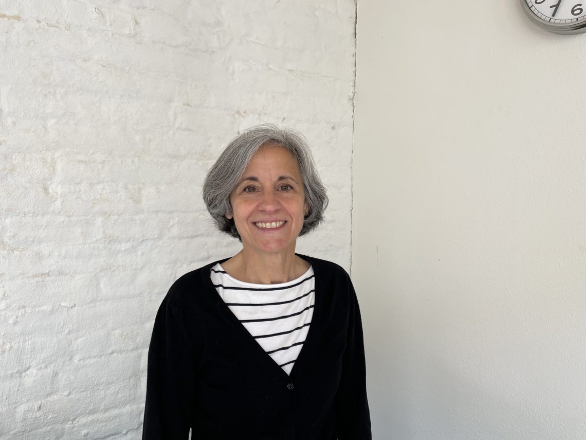 A person with short, gray hair is smiling at the camera. They are wearing a black cardigan over a white and black striped shirt. The background includes a white brick wall and a portion of a white clock on the top right corner, reminiscent of the Berkeley Journalism ambiance.