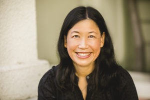 A woman with long black hair is smiling at the camera. She is wearing a black, long-sleeved top, and is sitting in front of a light-colored wall. The background is softly blurred, bringing focus to her bright and cheerful expression, reminiscent of candid moments often captured by Berkeley Journalism students.