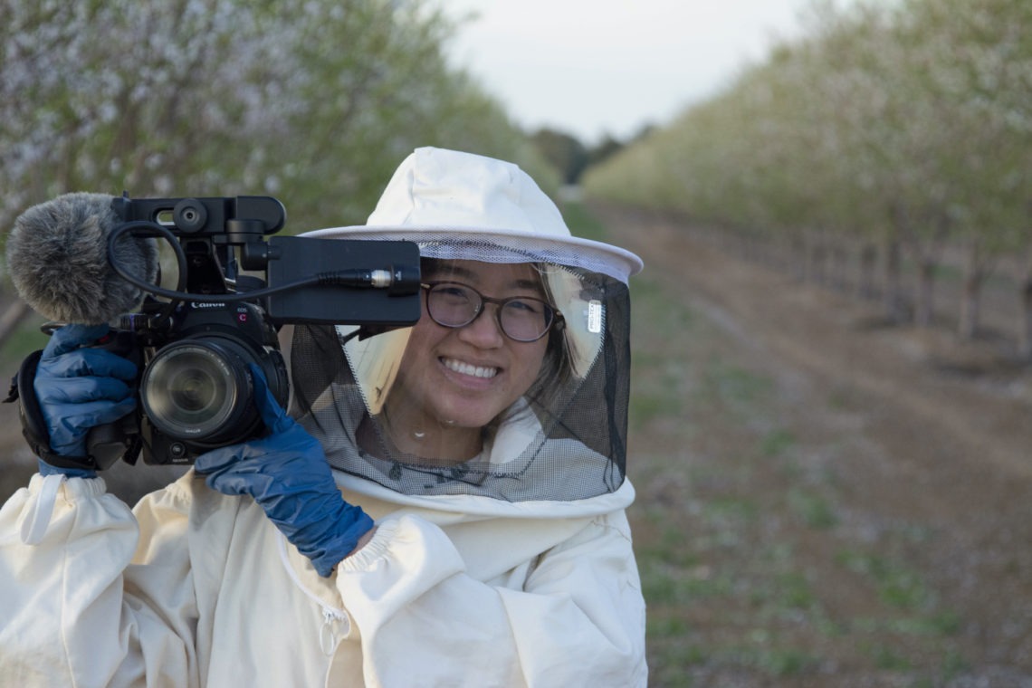 A young woman holding camera equipment outside while wearing a beekeeping suit.