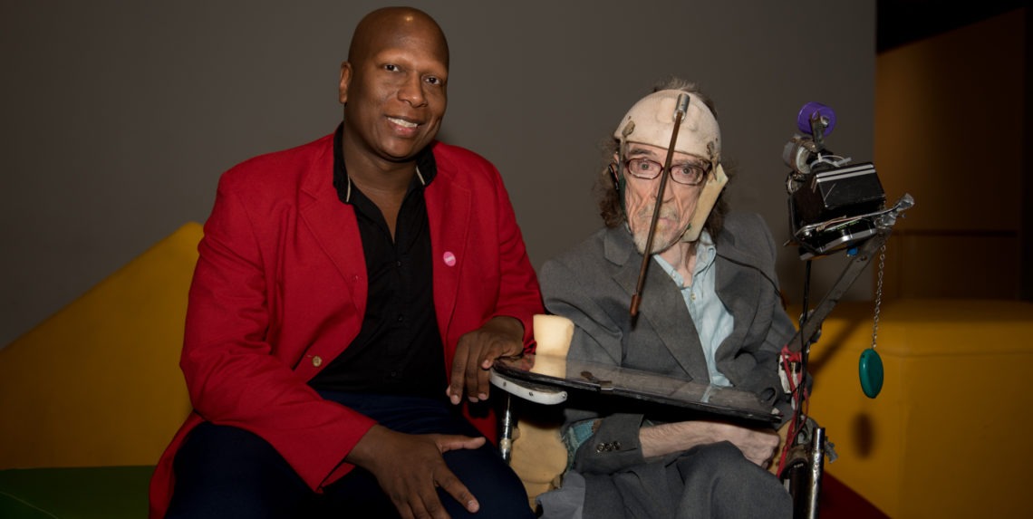 A person in a red blazer and another in a suit and hat, likely affiliated with Berkeley Journalism, sit together. The individual in the suit is using an assistive communication device mounted on their wheelchair. Both are looking at the camera and smiling against a simple, uncluttered background.