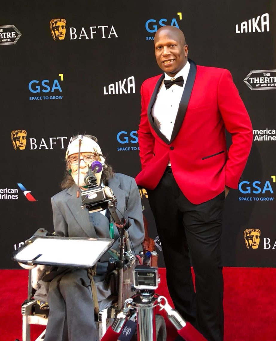 Two people at a formal event: one in a gray suit seated in a motorized wheelchair with medical equipment, the other standing and wearing a red tuxedo jacket with black lapels. They are on a red carpet with a backdrop featuring BAFTA and Berkeley Journalism sponsor logos.