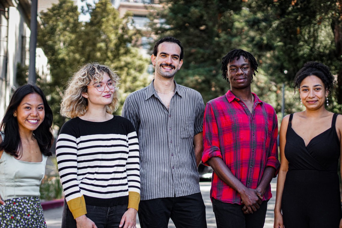 A group of five people standing outdoors and smiling at the camera. From left to right, a woman in a light green dress, a woman with curly blonde hair in a striped shirt, a man with a mustache in a gray Berkeley Journalism shirt, a man in a red plaid shirt, and a woman in a black dress.