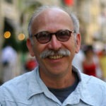 A bald man with glasses and a grey mustache is smiling at the camera. He is wearing a light blue shirt over a grey t-shirt. The background, reminiscent of Berkeley Journalism scenes, is blurred, showing hints of a city street with people and lights.