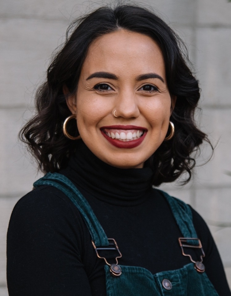 A smiling person with short, wavy dark hair, wearing hoop earrings, a black turtleneck, and green corduroy overalls stands confidently in front of a light gray brick wall. They have a nose piercing and an aura that captures the vibrant spirit of Berkeley Journalism.
