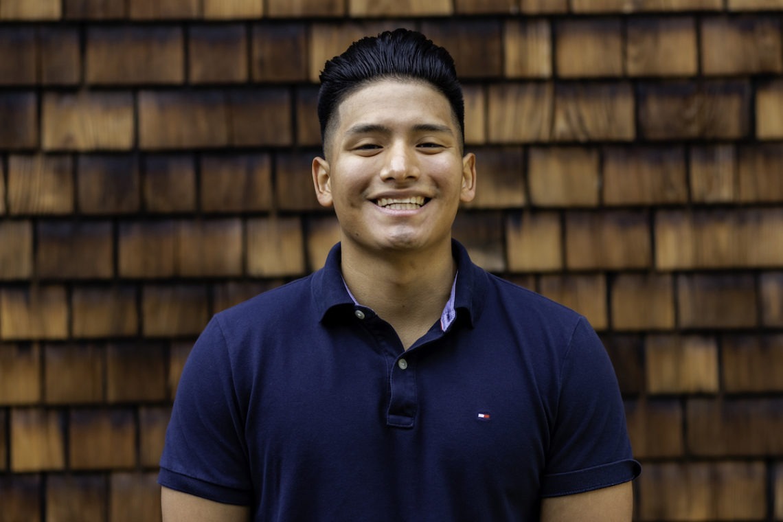 A smiling person with short, dark hair stands in front of a wooden shingle wall, wearing a navy blue polo shirt. Their confident demeanor hints at their background in Berkeley Journalism.