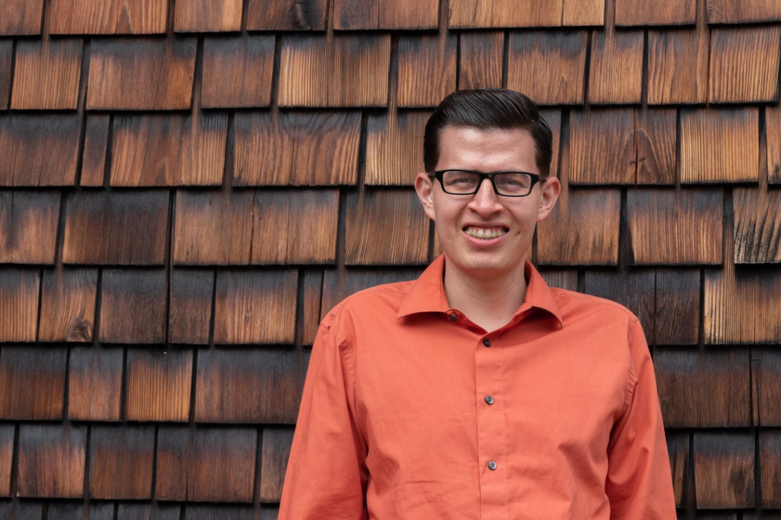 A man wearing black glasses and an orange button-up shirt stands smiling in front of a wooden shingle wall. The shingles, reminiscent of Berkeley Journalism's iconic settings, display a mix of light and dark brown tones, providing a textured backdrop.