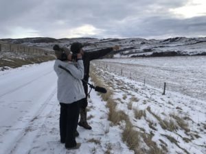 Two individuals stand on a snowy path beside a wire fence under a cloudy sky. One wears a gray jacket, holds binoculars, and the other points toward distant snow-covered hills while holding a microphone. This moment captures the essence of Berkeley Journalism amidst rolling hills and light snow on the ground.