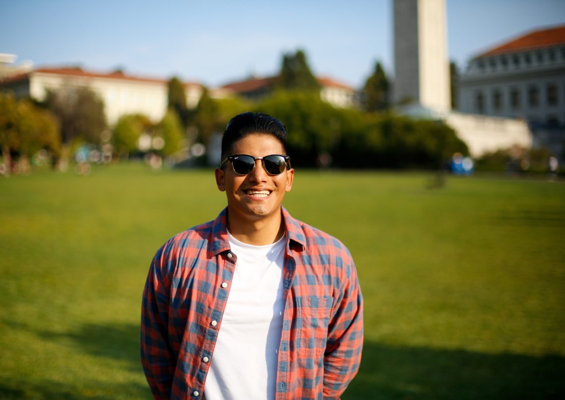 A man wearing sunglasses, a red and blue plaid shirt over a white T-shirt, smiles while standing in a sunlit grassy area with buildings and a tower in the background—perhaps capturing the essence of Berkeley Journalism.