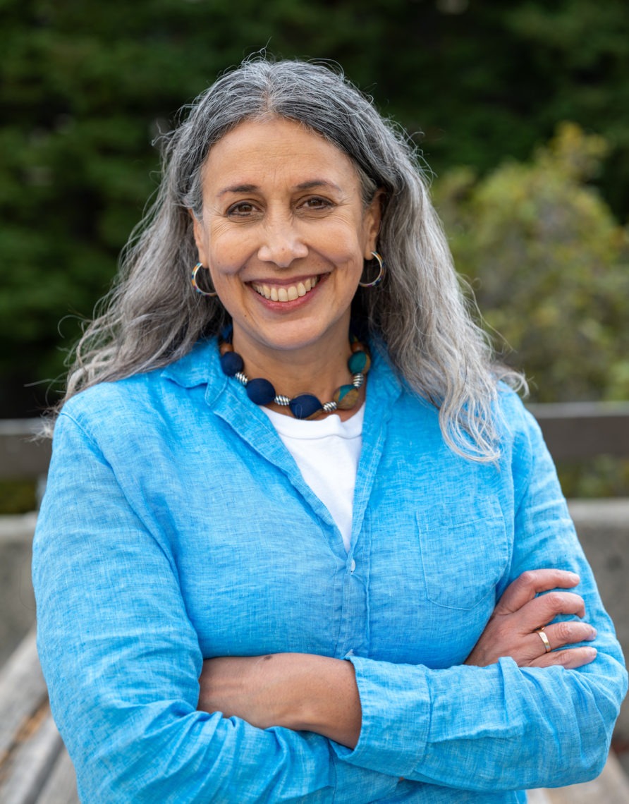 A woman with long, gray hair smiles at the camera with her arms crossed. She wears a light blue jacket over a white shirt and a colorful beaded necklace. The background features greenery with blurred trees and plants, capturing the thoughtful essence of Berkeley Journalism.