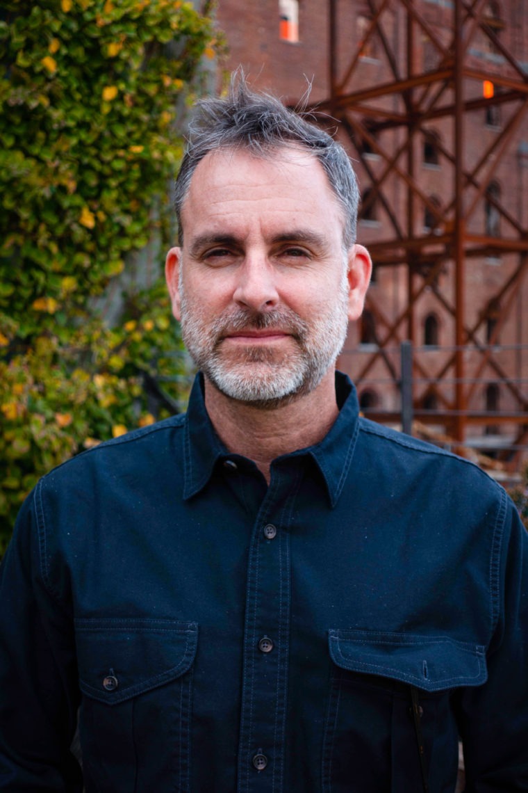A portrait of a middle aged man wearing a navy blue button up. He has short hair, a grey beard, and can be seen standing outside around a building with trees. 