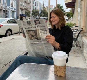 A woman sits at an outdoor table reading the San Francisco Chronicle newspaper. She is holding the newspaper with both hands, with a cup of coffee placed on the table in front of her. The street and residential buildings are visible in the background, evoking a sense of Berkeley journalism vibe.