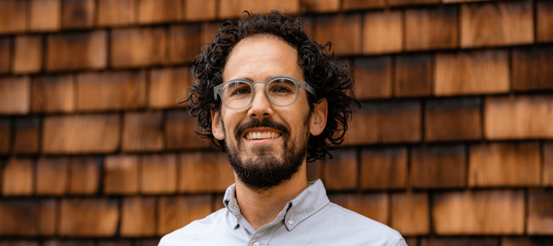 A man with curly hair and a beard, wearing glasses and a light-colored button-up shirt, smiles in front of a wooden shingle wall, exuding the thoughtful curiosity often found in Berkeley Journalism students.