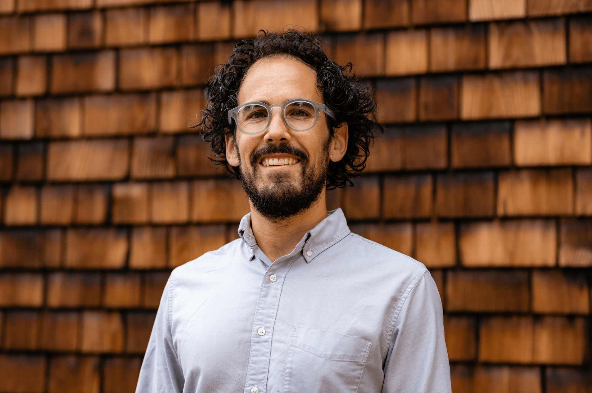 A person with curly hair, a beard, and glasses stands smiling in front of a wooden shingle wall. They are wearing a light gray button-up shirt, embodying the quintessential Berkeley Journalism vibe.