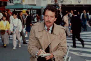 A man with a mustache wearing a beige trench coat and holding a notepad stands in focus amidst a bustling city crowd at a crosswalk, embodying the spirit of Berkeley Journalism. People around him are walking in various directions.
