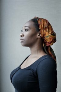 A young woman somberly looking off into the distance. She is wearing a head wrap and a black t-shirt.