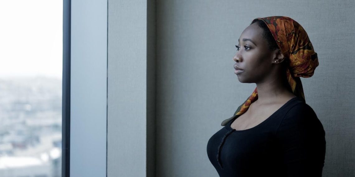 A woman dressed in a black top and an orange patterned headscarf is standing by a large window, gazing outside. Reminiscent of a Berkeley Journalism scene, the background is blurred, showing an out-of-focus cityscape. She appears thoughtful and serene.