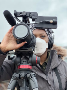 A woman using her camera and sound equipment outside. She is wearing a mask and a grey coat. 