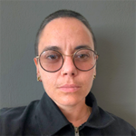 A person with close-cropped hair and round glasses looks directly at the camera with a neutral expression. They are wearing a black top. The background is plain and gray, reflecting the candid style often seen in Berkeley Journalism profiles.