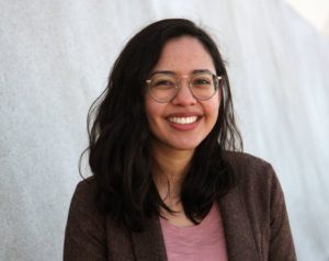A person with long, dark hair and wearing glasses smiles warmly at the camera. They are dressed in a brown blazer over a pink shirt, standing against a light-colored background, embodying the professional yet approachable spirit of Berkeley Journalism.