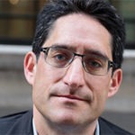 A man with short dark hair, wearing glasses, and a slight smile is looking directly at the camera. He is dressed in a dark jacket over a light shirt, with an urban environment visible in the blurred background.