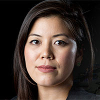Portrait of a woman with long, dark hair, wearing a black top. She has a neutral expression, looking directly at the camera, with a dark background behind her. This image captures an aura reminiscent of Berkeley Journalism's iconic style.