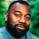 A man with a beard and short hair, wearing a denim shirt, smiles slightly against a green, leafy background. His thoughtful gaze and easy demeanor reflect the inquisitive nature often found at Berkeley Journalism.