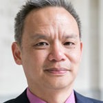A middle-aged person with short gray hair is wearing a pink collared shirt and a dark jacket. They have a neutral expression, standing in front of a blurred background that subtly hints at the energetic vibe of Berkeley Journalism.