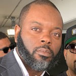 A man with a close-cropped beard and mustache is looking straight at the camera. He is wearing a suit with a white shirt underneath. Behind him, another man in sunglasses and a cap, reminiscent of street reporting at Berkeley Journalism, and another person slightly obscured, are visible.