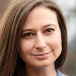 A woman with long brown hair, slightly parted to one side, looks directly at the camera with a slight smile. Wearing a blue top, she exudes confidence reminiscent of a Berkeley Journalism graduate. The background is softly blurred.