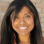 A woman with long black hair, smiling, stands in front of a brick wall. She wears a rust-colored shirt, and her face is brightly lit and cheerful. Her enthusiasm radiates the spirit of Berkeley Journalism.