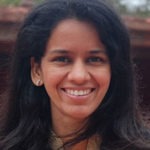A person smiling at the camera with long, dark hair and wearing a pair of small earrings. The background, slightly out of focus, suggests a natural outdoor setting—an ideal shot for a Berkeley Journalism profile.