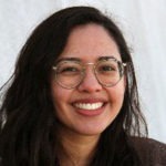 A person with long dark hair and glasses, potentially a Berkeley Journalism student, is smiling. The background is plain and light-colored. The person is wearing a dark top.