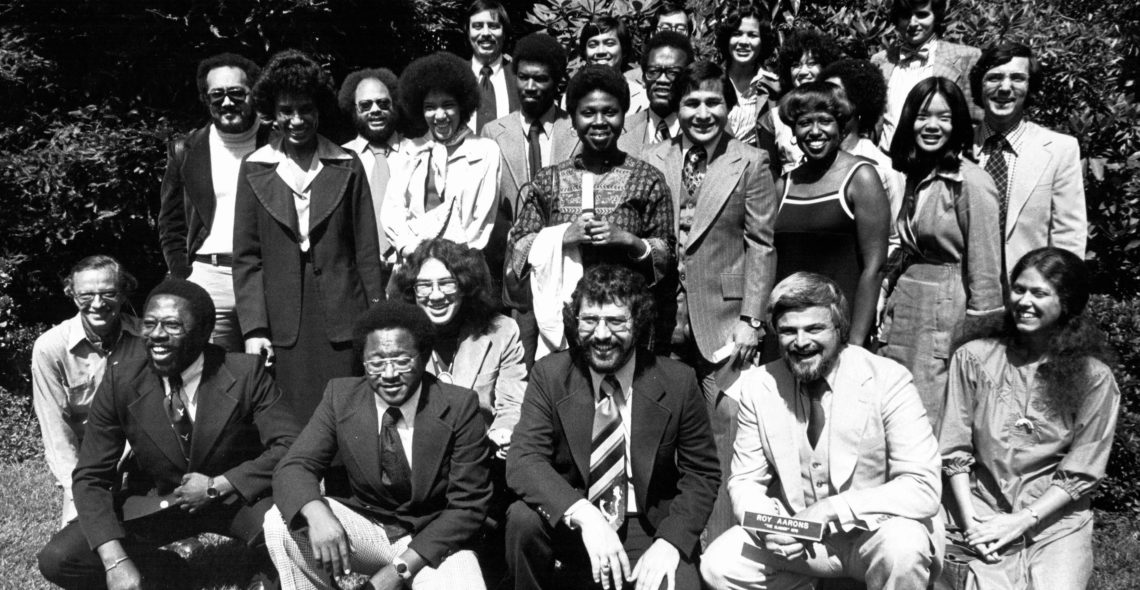 A black and white photo of a diverse group of twenty-three people, including both men and women, posed outdoors. The individuals, perhaps part of a Berkeley Journalism cohort, are dressed in 1970s style clothing, with many smiling at the camera. Trees and foliage are visible in the background.
