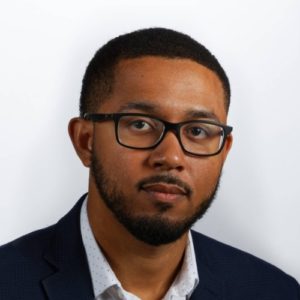 A man with short black hair, a beard, and glasses is wearing a white dress shirt and a dark blazer. He is looking directly at the camera with a neutral expression, set against a plain white background, embodying the professionalism often associated with Berkeley Journalism.