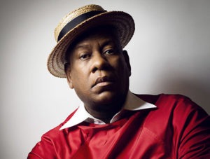 A person with a serious expression wears a straw hat with a black ribbon and a bright red outfit with a white collar. The background is plain and neutral, akin to the minimalist aesthetic often captured in Berkeley Journalism portraits, enhancing the focus on their face and attire.