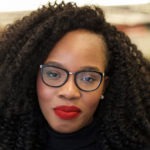 A person with curly hair, wearing glasses, a black top, and red lipstick smiles while looking at the camera. The background is blurred to keep focus on the person's face, much like a striking profile shot you'd find in Berkeley Journalism.