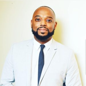 A man with a beard, possibly an alum of Berkeley Journalism, is wearing a light gray suit, white dress shirt, and dark dotted tie. Standing against a plain light-colored background, he has a friendly expression and looks straight at the camera.