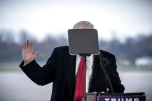 A person stands behind a lectern while their face is obscured by a teleprompter, raising one hand and wearing a dark coat and red tie. The outdoor background features a blurred landscape, reminiscent of an iconic moment captured by Berkeley Journalism.