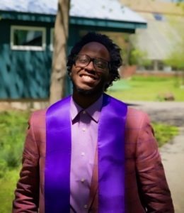 A man smiles broadly while standing outside on a sunny day. He's wearing glasses, a light purple shirt, a pinkish patterned jacket, and a bright purple graduation stole from Berkeley Journalism. Green trees and a building with teal siding are visible in the background.