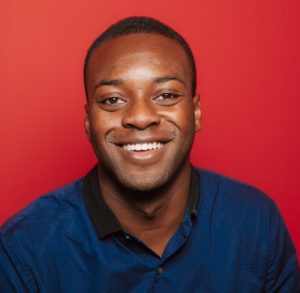 A man with short hair, wearing a blue shirt with a black collar, smiles in front of a red background, embodying the vibrant spirit of Berkeley Journalism.