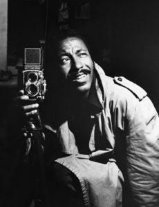 Black and white photo of a man wearing a trench coat, looking upwards while holding a vintage Rolleiflex camera. The image captures the essence of timeless curiosity and pays homage to the spirit of Berkeley Journalism.