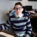 A person with short hair, wearing black-framed glasses, a striped sweater, and a necktie sits at a desk, smiling while looking towards the camera. A partially visible laptop and blurred office background hint at their productive day in the Berkeley Journalism department.