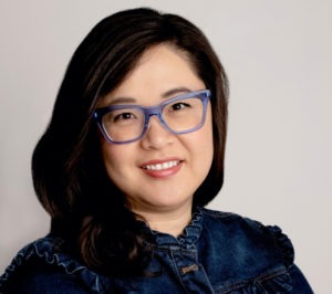 A woman with long dark hair is wearing blue-framed glasses and a dark blue, ruffled denim top. She is smiling and posed against a plain background, embodying the vibrant spirit of Berkeley Journalism.