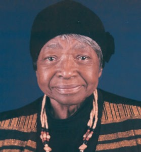 An older person with a warm smile wears a black hat and a black outfit with gold patterns, reminiscent of the sophisticated style often seen in Berkeley Journalism circles. They also have a necklace made of beige and brown beads. The background is a solid blue color.