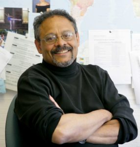 A man with glasses and a goatee is sitting with his arms crossed, smiling at the camera. He is wearing a black turtleneck and is in an office adorned with various papers, maps, and documents pinned to a bulletin board behind him, reflecting the investigative spirit of Berkeley Journalism.