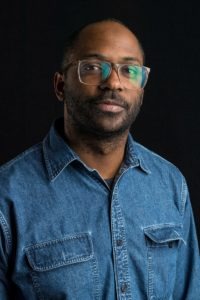 A man with short hair and a beard wearing glasses and a denim shirt stands against a black background. With a neutral expression, he gazes slightly to his right, embodying the thoughtful demeanor often associated with Berkeley Journalism.