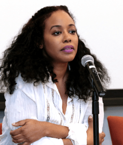 A person with long, curly hair and wearing a white blouse speaks into a microphone. They have a serious expression and their arms are crossed, embodying the focused determination associated with Berkeley Journalism. The background is plain and the image is taken indoors.
