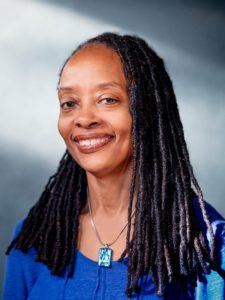 A woman with long, black dreadlocks smiles at the camera. She is wearing a blue top and a necklace with a blue pendant. The background is softly lit and blurred, highlighting her features, reminiscent of the vibrant portraits often seen in Berkeley Journalism pieces.