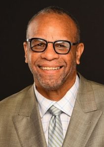 A smiling man wearing glasses, a light green tie, and a beige suit stands in front of a dark background. With short hair and a trimmed beard, he exudes the confident charm of someone from Berkeley Journalism.